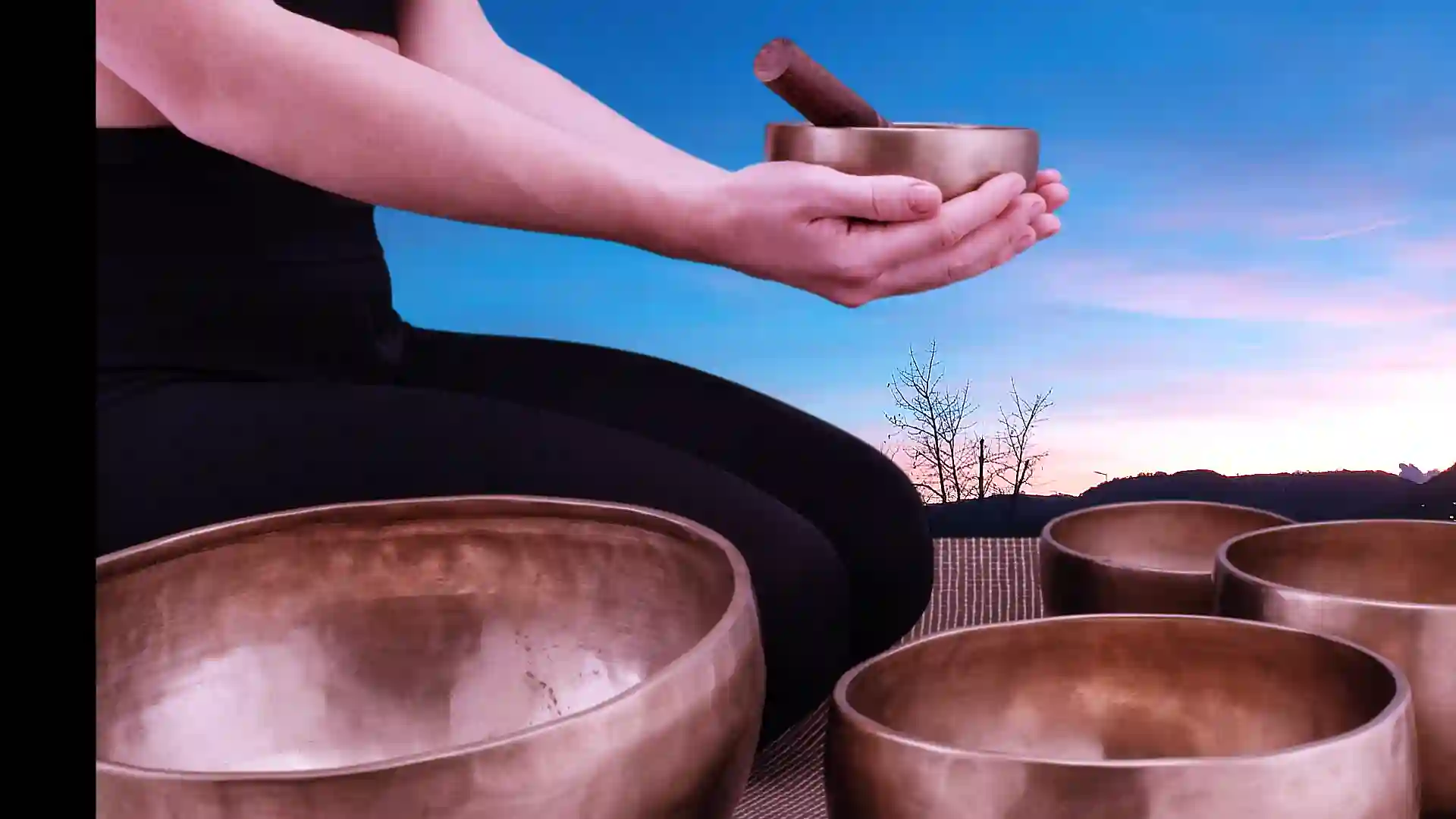 Bols tibétains tenus par des mains dans un décor naturel au coucher du soleil pour séance de bain sonore